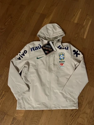 Vit Nike Brasil regnjacka GORE-TEX - Snygg vit regnjacka från Nike med Brasilien-logga och sponsormärken. Jackan har huva, hel dragkedja och är tillverkad i vattentätt GORE-TEX-material. Perfekt för dig som gillar fotboll och vill hålla dig torr med stil.