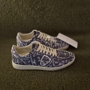 Philippe Model skor - Snygga sneakers från Philippe Model med marinblå bas och vitt bandanamönster över hela skon. Skorna har vita snören, rund tå och platt sula. Perfekta för dig som vill sticka ut med en cool och unik design. Materialet är skinn och textil.