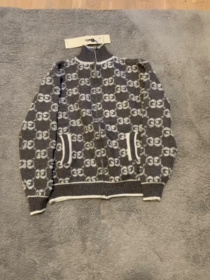 Grå mönstrad kofta från Gucci - Säljer en grå och vit stickad kofta från Gucci med GG-logga över hela plagget. Koftan har dragkedja framtill, ribbad krage och muddar samt två fickor med vit kant. Perfekt för dig som gillar lyxiga och snygga detaljer. Storlek S 
