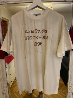 Vit Acne Studios t-shirt med strass - Vit oversized t-shirt från Acne Studios med tryck 'Acne Studios STOCKHOLM 1996' framtill och små strassdetaljer över hela plagget. Klassisk rund hals och kort ärm. Tillverkad i mjuk bomull för en skön känsla och clean look.