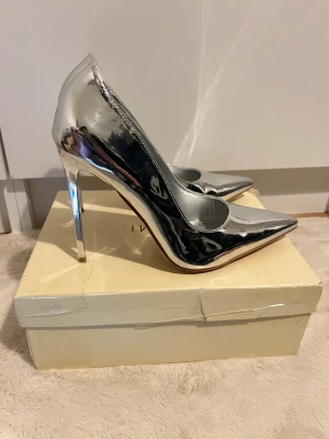 Simmi London stilettklackar - Säljer ett par riktigt snygga klackar från Simmi London i metallic silver. Skorna har spetsig tå, hög klack och en glansig finish som verkligen sticker ut. Perfekta för dig som vill ha ett statement-piece till outfiten. Materialet är syntet och de har en klassisk, elegant form. Den är 12 cm hög.