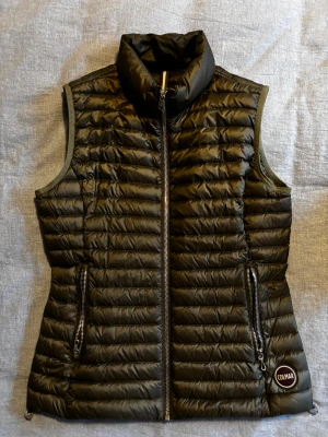 Mörkgrön dunväst från Colmar - Snygg svart dunväst från Colmar med hög krage. Västen har två dragkedjeförsedda fickor och Colmar-logga på framsidan. Tillverkad i glansig polyester och dunfyllning, perfekt för lager-på-lager. Stängs med dragkedja framtill. Helt oanvänd