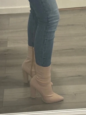 Beige stövletter med klack och spetsig tå - Säljer ett par snygga beige stövletter med hög klack och spetsig tå. Skorna har dragkedja på insidan och är gjorda i ett stretchigt syntetmaterial som sitter tight runt vristen. Perfekta för dig som vill ha en trendig och stilren look.