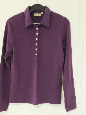 Lila tröja med knappar och krage - En lila långärmad tröja med krage och knappar framtill. Tillverkad i mjuk bomull (92 %) och elastisk elastan (8 %). Perfekt för dig som gillar stilrena plagg. Tröjan har en naturligt tvättad look. Se bilder för skick och passform. Färgen på bilderna, speciellt närbilderna, stämmer inte alltid överens med verkligheten. Storlek M enligt lappen men då måttet armhåla till armhåla är cirka 43 cm så stämmer S bättre. Axelsöm till ärmslut är cirka 60 cm. Tvättas i 60 grader.