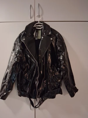 Svart vegan skinnjacka Divided S - Säljer en svart, glansig vegan skinnjacka från Divided i storlek S. Jackan har coola detaljer som snedställd dragkedja, bred krage och bälte nedtill. Perfekt för dig som vill ha en edgy look med mycket attityd.