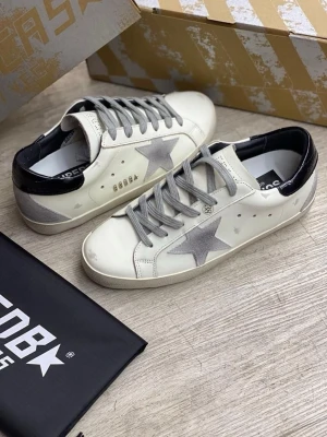 Golden Goose vita sneakers med stjärna - Snygga vita sneakers från Golden Goose med grå mockadetaljer och en ikonisk stjärna på sidan. Skorna har grå snörning, mörkblå häl och platt sula. Perfekta för dig som gillar en cool och avslappnad stil med unika detaljer.ps boxen medföljer 