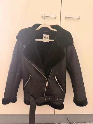 Svart pälsjacka från Zara - Snygg svart pälsjacka från Zara med bred krage, dragkedja i sned modell och två framfickor med dragkedjor. Jackan har bälte i midjan och mjukt foder som syns vid ärmslut och nederkant. Perfekt för kalla dagar och ger en cool look.