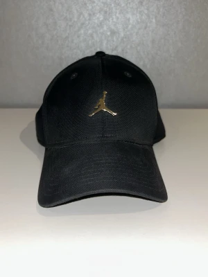 Svart Jordan keps med gulddetalj - Snygg svart keps från Jordan med det ikoniska Jumpman-loggan i guld framtill. Klassisk böjd skärm och justerbar passform. Tillverkad i slitstarkt tyg som ger en clean och sportig look. Perfekt accessoar för dig som gillar streetwear och sportmode.