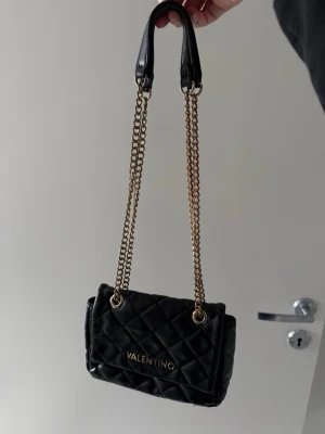 Svart valentino väska - Svart valentino väska med guldiga kedjor på handtagen. Väskan har slitningar på lädret samt kedjorna som man kan se på bilderna ovanför.