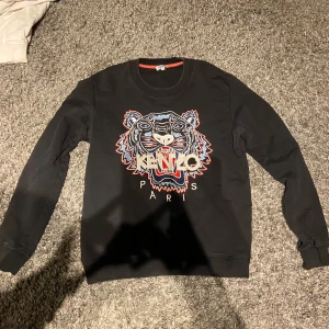 Svart Kenzo sweatshirt med tigertryck - Cool svart sweatshirt från Kenzo med ikoniskt broderat tigerhuvud och texten 'KENZO PARIS' på bröstet. Tröjan har rund halsringning, ribbade muddar och är perfekt för dig som gillar streetwear och statement-plagg.