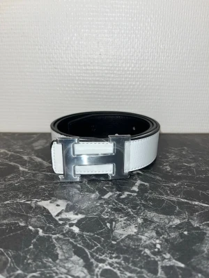 Vändbart skärp i skinn - Stilrent vändbart skärp i svart och vitt skinn med ikoniskt H-spänne i silver. Perfekt accessoar för att lyfta din outfit med en lyxig touch. Bältet har en klassisk design och passar till många olika stilar.