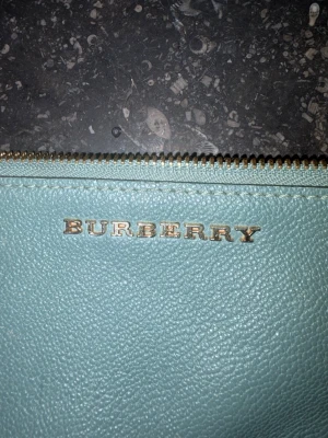 Grön axelväska från Burberry - Stilren grön/blå axelväska från Burberry i läder med guldfärgade detaljer och justerbar axelrem med kedja. Väskan har dragkedja upptill och klassiskt rutigt foder samt inbyggda kortfack. Perfekt för dig som gillar lyxiga accessoarer med smarta detaljer.väskan är såklart äkta köpt från Heathrow flygplats i london.