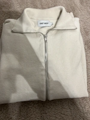 Vit ziptröja från EAST WEST - Säljer en stilren vit ziptröja från EAST WEST. Tröjan har ribbad struktur, hög krage och dragkedja framtill. Perfekt för lager-på-lager och enkel att matcha med andra plagg. Materialet känns mjukt och bekvämt.priset kan diskuteras vid snabb affär 🫱🏻‍🫲🏽