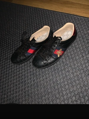 Gucci Ace svarta skor med bi - Svarta Gucci Ace sneakers i läder med klassiska röd-blå ränder på sidan och broderad guldfärgad bi. Röd metallic-detalj på hälen och mönstrad innersula. Snygg och tidlös modell med svarta skosnören.
