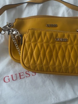 Gul handväska från Guess - Två snygga handväskor från Guess i en härlig gul färg. Ena väskan är quiltad med diamantmönster och silverdetaljer, den andra är slät med dragkedja. Båda har Guess-logga i metall och justerbara axelremmar, en i läder och en i kedja. Perfekt för att lyfta din outfit.