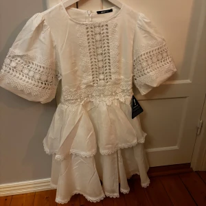 Vit volangklänning med spetsdetaljer - Superfin vit kortklänning från ginateicot  med volanger och spetsdetaljer. Klänningen har puffiga kortärmar med broderade spetsinslag och vacker spets framtill. Perfekt för sommarens alla tillfällen! Köpt för 800kr men säljer för 650, aldrig använd, prislapp kvar. Säljer så den ej passar