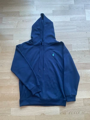 Marinblå hoodie från Polo Ralph Lauren - Klassisk marinblå hoodie från Polo Ralph Lauren med broderad grön logga på bröstet. Hoodien har stor huva med dragsko, känguruficka och ribbade muddar vid ärmar och nederkant. Tillverkad i mjuk bomullsblandning för extra komfort.