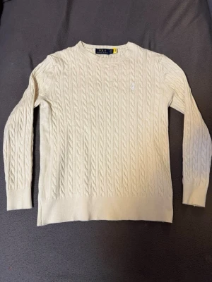 Beige kabelstickad tröja från Polo Ralph Lauren - Kabelstickad vit tröja från Polo Ralph Lauren med rund halsringning och diskret broderad logga på bröstet. Tröjan har långa ärmar och ribbade muddar vid ärmslut och nederkant. Perfekt för en stilren och klassisk look.