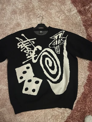 Svart stickad tröja från Stüssy - Säljer en svart stickad tröja från Stüssy med vita grafiska mönster, bland annat tärningar, spiral och graffiti-liknande detaljer. Tröjan har rund halsringning och ribbade muddar. Perfekt för dig som gillar streetwear och unika plagg.