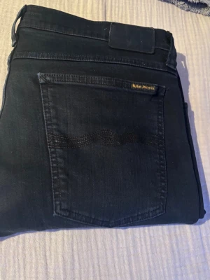 Svarta jeans från Nudie Jeans - Säljer ett par svarta jeans från Nudie Jeans med klassisk femficksmodell och diskret vågmönster på bakfickan. Jeansen har en smal passform och är tillverkade i mjuk denim med en liten etikett på fickan. Perfekta för en stilren och avslappnad look. (W34, L32)