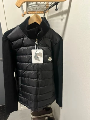 Svart cardiganjacka från Moncler - Snygg svart cardiganjacka från Moncler med quiltad front i glansigt material och stickade ärmar och rygg. Jackan har dragkedja framtill och Moncler-logga på bröstet. Perfekt för lager-på-lager och en stilren look.