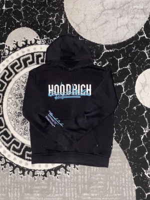 Svart HOODRICH hoodie med blått tryck - Svart hoodie från HOODRICH med vit och blå text på bröstet och ärmen samt stort blått tryck på ryggen. Klassisk modell med huva och dragsnören. Perfekt för en streetwear-look. Mjuk bomullskänsla och avslappnad passform.