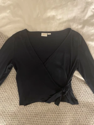 Svart omlottopp från Gina Tricot -  Mörkblå tröja från Gina Tricot med långa ärmar och knyt i sidan. Toppen har en djup v-ringning och är tillverkad i ett mjukt, stretchigt material. Storlek 170. 