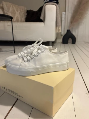 Vita skor - Klassiska vita sneakers i canvas med lågt skaft och vita snören. Skorna har en vit gummisula med diskreta linjer och förstärkt tåparti. Perfekta för en clean och enkel stil.