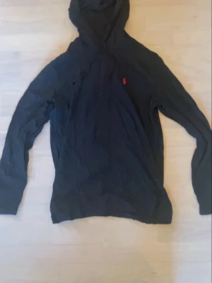 Marinblå hoodie från Ralph Lauren - Snygg marinblå hoodie från Ralph Lauren med klassisk röd logga broderad på bröstet. Hoodien har huva med dragsko och är tillverkad i mjukt material som känns skönt mot huden. Perfekt för en avslappnad och stilren look.