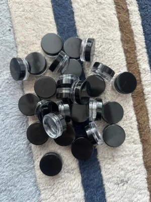 Svarta tomma burkar för nageldekoration - Ett gäng små, runda plastburkar med svarta lock, perfekta för att förvara nageldekorationer som glitter, pärlor eller stenar. Burkarna är genomskinliga på sidorna så du ser enkelt innehållet. Smidiga att ta med och hålla ordning på dina nagelgrejer. Finns lite pärlor i några som man får med gratis