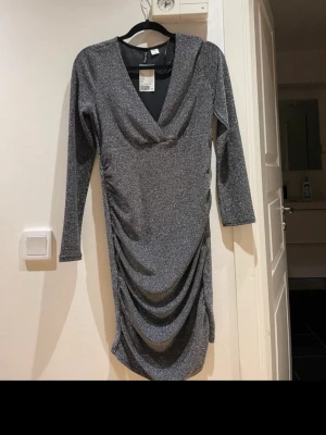 Glittrig silverklänning med v-ringning - Snygg glittrig silverfärgad klänning från H&M med v-ringad hals, långa ärmar och draperad effekt framtill. Kort modell som sitter snyggt på kroppen och passar perfekt när du vill sticka ut lite extra.