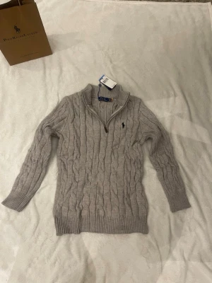 Grå kabelstickad tröja Polo Ralph Lauren - Snygg grå kabelstickad tröja från Polo Ralph Lauren med halv dragkedja och hög krage. Tröjan har långa ärmar, ribbade muddar och den klassiska loggan broderad på bröstet. Perfekt för en stilren och avslappnad look.