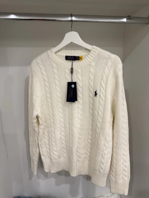 Vit  tröja från Polo Ralph Lauren - Kabelstickad vit tröja från Polo Ralph Lauren med rund halsringning och klassisk logga broderad på bröstet. Tröjan har långa ärmar och ribbade muddar vid ärmslut och nederkant. Perfekt för en stilren och tidlös look.