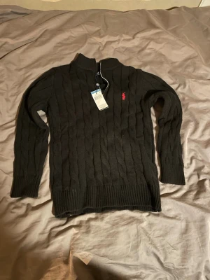 Svart kabelstickad tröja Polo Ralph Lauren - Kabelstickad svart tröja från Polo Ralph Lauren med halv dragkedja och röd broderad logga på bröstet. Tröjan har ribbade muddar och hög krage, perfekt för en stilren och klassisk look.