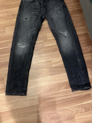 Svarta Jack&jones jeans med slitningar - Säljer ett par svarta jeans med coola slitningar och tvättade detaljer. Jeansen har rak passform och klassisk femficksdesign. Perfekta för en avslappnad och trendig look. Storlek 29w 32l
