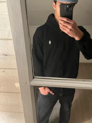 Svart hoodie/zip tröja från Ralph Lauren - Svart hoodie med dragkedja/zip up tröja från Ralph Lauren. Klassisk design med vit broderad logga på bröstet, huva med dragsko och känguruficka framtill. Mjuk bomullskänsla och ribbade muddar vid ärmslut och nederkant. Perfekt för en chill och stilren look. Storlek L fast sitter som S!
