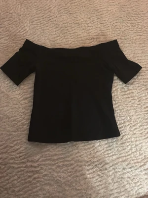 Svart offshoulder topp - Snygg svart offshoulder topp med korta ärmar. Enkel och stilren design som passar perfekt till jeans eller kjol. Mjuk och stretchig känsla i materialet för en bekväm passform.
