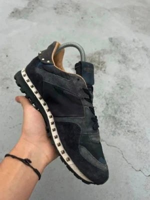 Valentino skor - Snygga svarta sneakers från Valentino med coola nitar längs sulan och detaljer i mocka och textil. Skon har en låg profil, svarta skosnören och camo-mönster på vissa partier. Perfekt för dig som vill sticka ut med en exklusiv touch. Priset kan diskuteras