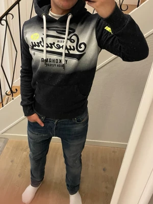 Superdry hoodie med tryck, svart/grå - Snygg hoodie från Superdry i svart och grått med stort tryck på bröstet där det står 'Superdry Yokohama High Flyers'. Hoodien har vit dragsko i huvan och en klassisk känguruficka framtill. Perfekt för en avslappnad streetwear-look.