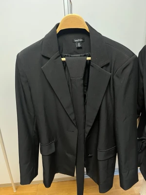 Svart kavaj från Boohoo - Stilren svart kavaj från Boohoo med klassisk krage och två fickor framtill. Kavajen har en rak passform och är helfodrad för extra komfort. Perfekt för att lyfta vilken outfit som helst med en clean och modern look.