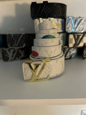 Vitt Läder bälte med guldspänne - Snyggt vitt läder bälte med ikoniskt mönster och stort guldfärgat spänne. Bältet har färgglada detaljer och en gul etikett med logotypen på baksidan. Perfekt accessoar för att lyfta din outfit.