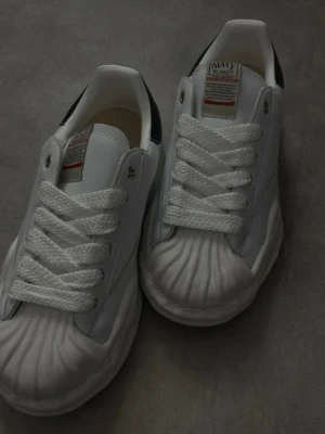 Maison Margiela  - Unika vita sneakers från maison margiela med tydliga snäckformade detaljer på tån och tjock vit sula. Skorna har svarta detaljer vid hälen och guldtext på sidan. Klassisk låg modell med vita skosnören och robust design. Perfekta för dig som gillar statement sneakers.