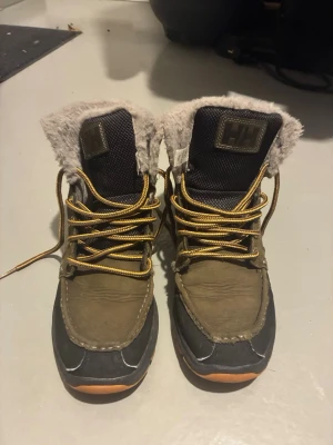 Helly Hansen fodrade vinterkängor - Snygga vinterkängor från Helly Hansen med grått foder, bruna och svarta partier samt gula snören. Kängorna har en robust sula och är perfekta för kalla dagar. Fodret är mjukt och värmande, och HH-loggan syns tydligt både fram och bak. Andvända cirka 5 gånger i snön så de är som helt nya ordinarie pris 2000kr ish!