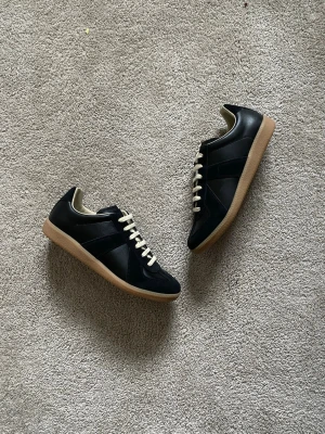 Maison Margiela Gats Svarta - Stilrena svarta  Maison Margiela sneakers med beige gummisula och vita snören. Skorna har en klassisk låg profil och är tillverkade i en mix av skinn och mocka. Perfekta för dig som gillar minimalistisk och tidlös design.