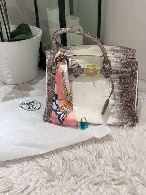 krokodilskinn handväska - Exklusiv handväska i krokodilskinn med ljusbeige mitt och gråbruna kanter. Väskan har guldfärgat lås, handtag och en färgglad scarfdetalj i rosa och orange. Perfekt för dig som vill sticka ut med lyxig design.