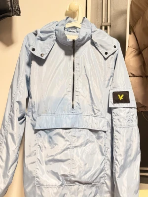 Ljusblå vindjacka från Lyle & Scott - Snygg ljusblå vindjacka från Lyle & Scott med halv dragkedja framtill, stor ficka på magen och huva. Jackan har en patch med gul logga på ärmen och är tillverkad i glansigt, lätt material. Perfekt för blåsiga dagar.