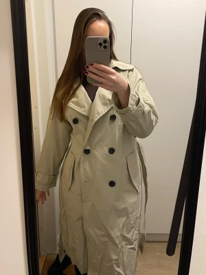 Beige klassisk trenchcoat med knappar - Säljer denna stilrena trenchcoaten med dubbelknäppning och stora svarta knappar. Lång modell med bred krage och två snedställda fickor framtill. Bälte tillkommer! 