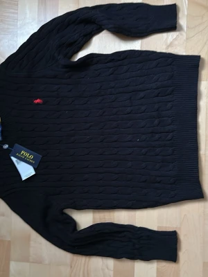 Svart kabelstickad tröja Polo Ralph Lauren - Kabelstickad svart tröja från Polo Ralph Lauren med den klassiska röda loggan broderad på bröstet. Tröjan har långa ärmar, ribbade muddar och rund hals. Perfekt för dig som gillar stilrena och tidlösa plagg. PRIS KAN DISKUTERAS SKRIV VID EVENTUELLA FRÅGOR (använd gärna Bring som leverantör)