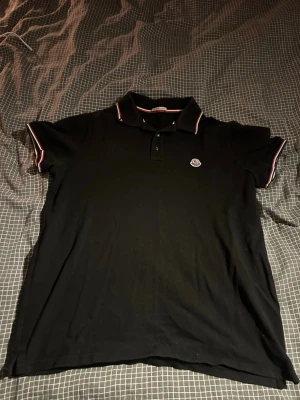 Svart Moncler pikétröja - Klassisk svart pikétröja från Moncler med röd och vit kant på krage och ärmslut. Tröjan har korta ärmar, knappslå framtill och Moncler-logga på bröstet. Perfekt till en clean och stilren look. Storleken är XL men passar till en L.