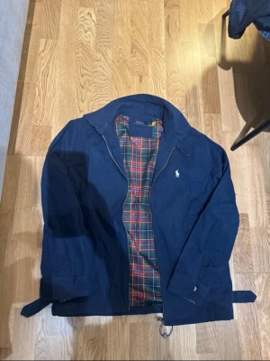  Ralph Lauren harringtonjacka - Stilren marinblå vindjacka från Polo Ralph Lauren med dragkedja och klassisk vit logga på bröstet. Insidan har ett färgglatt rutigt foder i rött, grönt och gult. Jackan har hög krage och justerbara ärmslut för extra komfort.